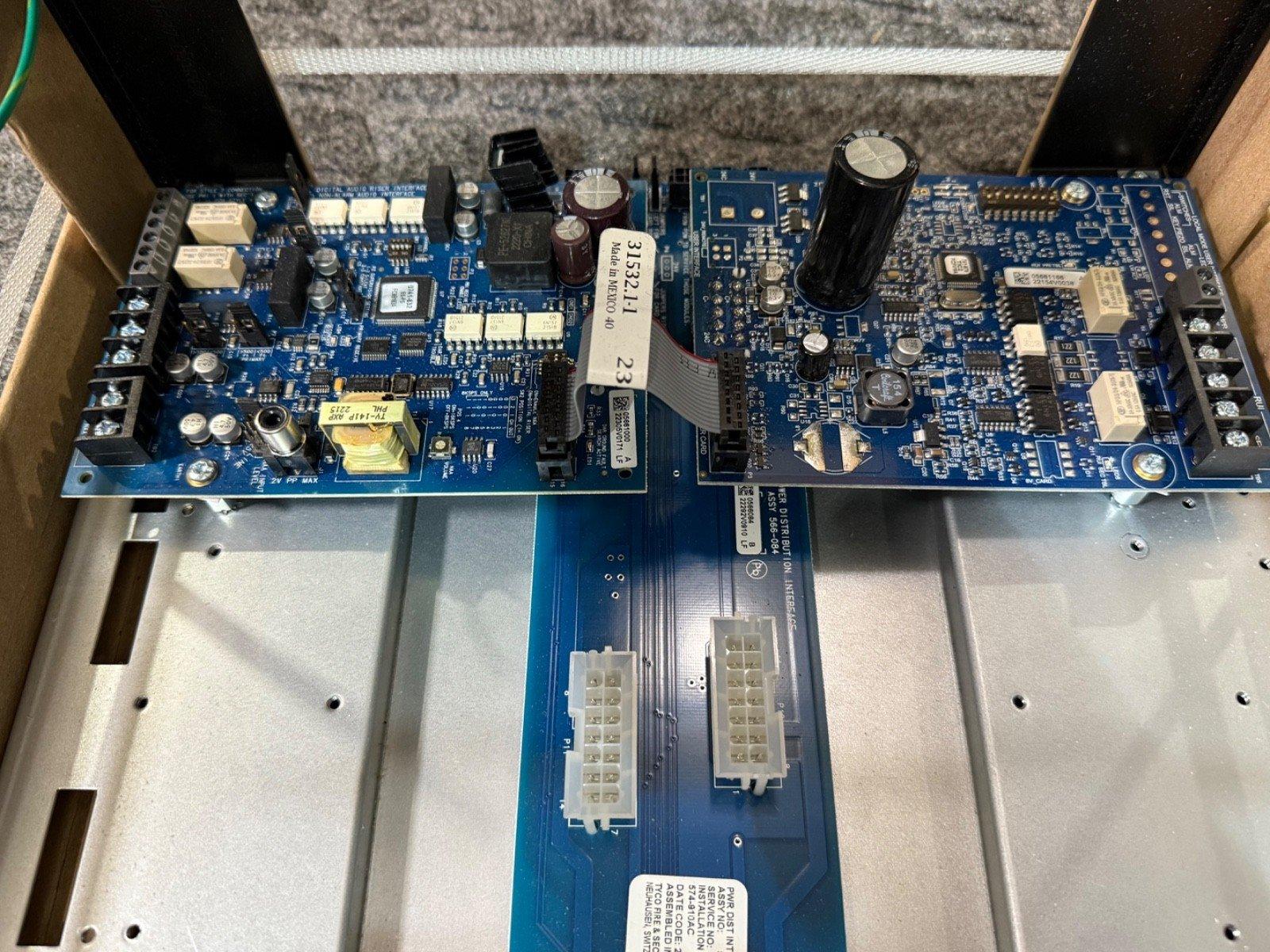 Blue fire alarm control panel circuit board with capacitors, connectors, and labels inside metal enclosure