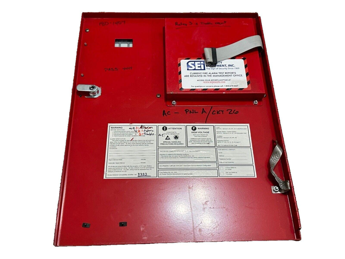 Red fire alarm control panel with SEI label, warning stickers, and handwritten notes for maintenance and testing.