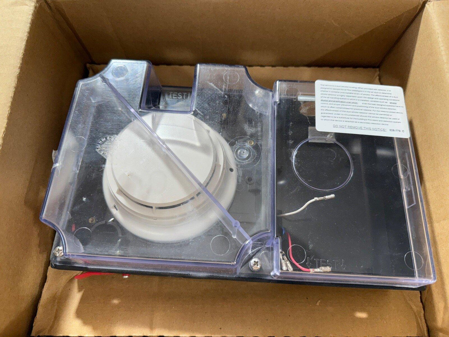 Fire alarm smoke detector in clear housing with test button, wiring, and warning label inside box